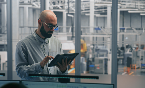 A bald, bearded man wearing glasses and a grey collared shirt uses a tablet in a factory control room. He is focused on the screen in front of him. The background shows industrial machinery and production lines.